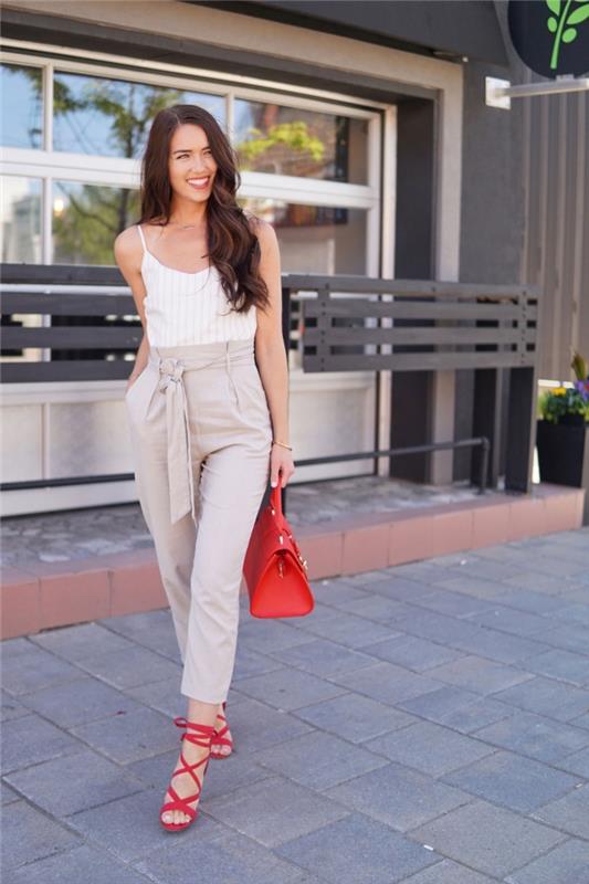 dobro oblečena ženska v priložnostnem elegantnem kombinezonu z ozkimi hlačami v bež barvi in ​​črtastim vrhom v beli barvi, opremljena s sandali in rdečo torbico