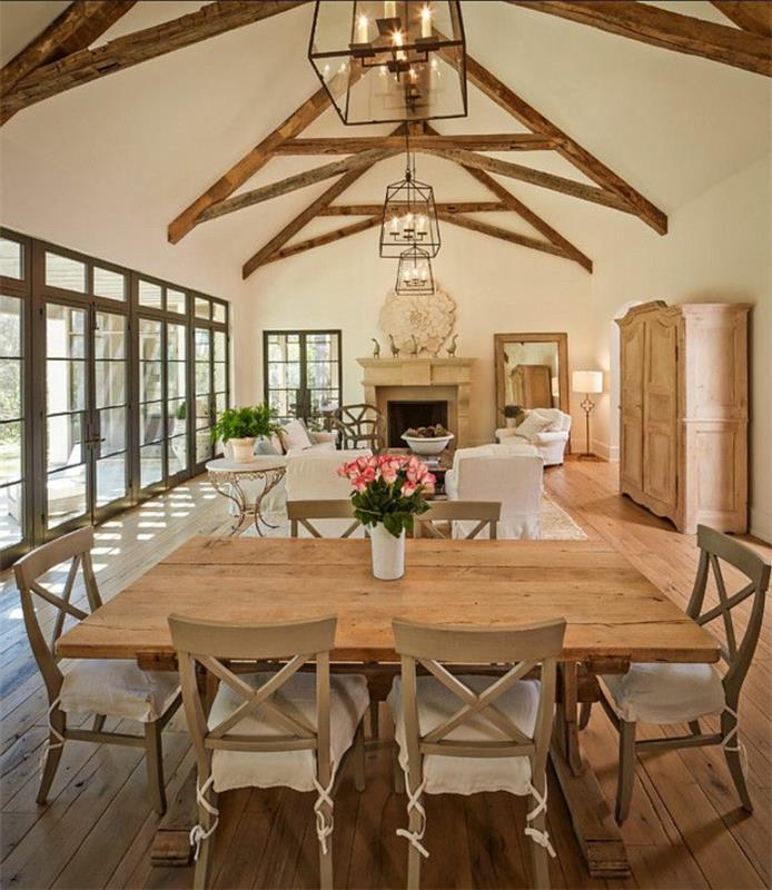 eleganten podeželski dekor, lesena jedilna miza in stoli, izpostavljeni tramovi, beli naslanjači, lesena tla, viseče luči, kamniti kamin