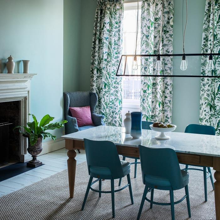 valgomasis akvareliniais dažais, akmeninis židinys, medinis stalas su marmurine viršuje, senovinės kėdės iš ančių mėlynos spalvos metalo, baltos užuolaidos su žalių lapų raštais, pilkas kilimas