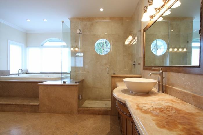 Baltas stalviršio praustuvas, įmantrus marmurinis stalviršis, apšviestas veidrodis ir didelė pakelta vonia, travertino vonios kambarys