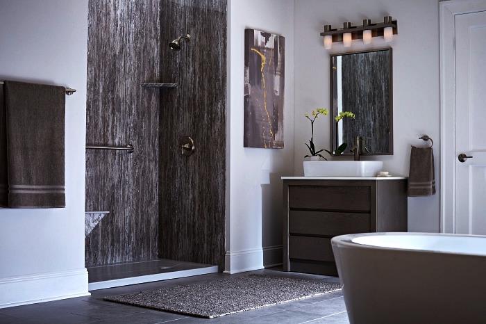 Zen in model naravne kopalnice v beli in sivi barvi s tušem, ki se odpira v prostor s stenami z učinkom naravnega kamna in kadjo sive barve