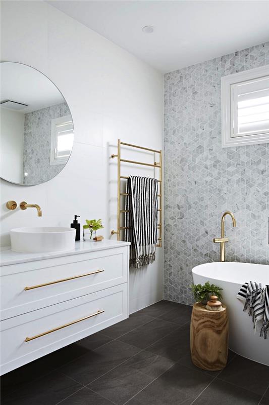 sivo-bela kopalnica s čistimi oblogami s sodobno samostoječo kadjo in poudarjeno steno s šestkotnimi ploščicami v odtenkih sive, pipami iz medenine in ogrevanimi brisačami
