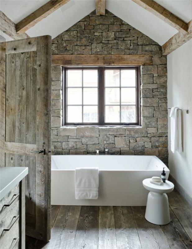 Dekoracija kopalnice na podeželju, kamniti zid, parket in vrata iz surovega lesa, samostoječa kad, izpostavljeni tramovi, bele brisače