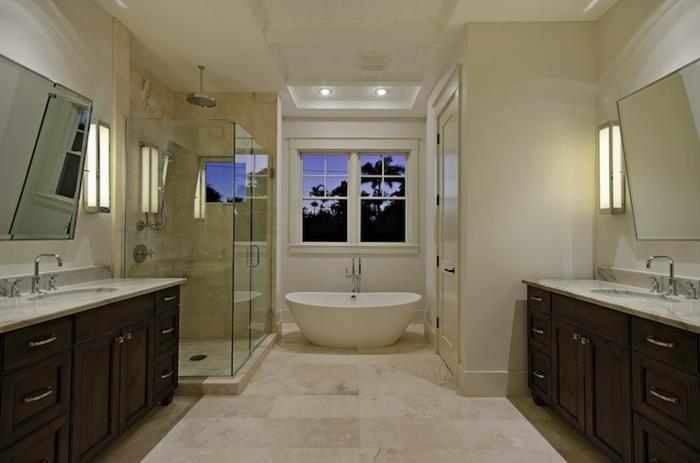 kreminės spalvos vonios kambarys, balta vonia po dideliu langu, travertino plytelės