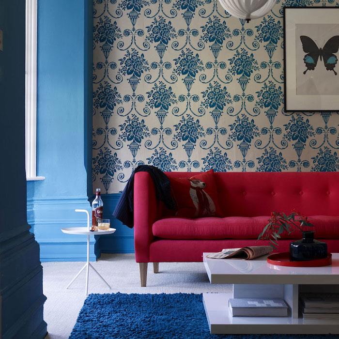 mėlyna ir raudona svetainė, spalvų schema, balti tapetai su mėlynais gėlių raštais, mėlynas kilimas ir baltas kavos staliukas, raudona sofa, senovinė atmosfera