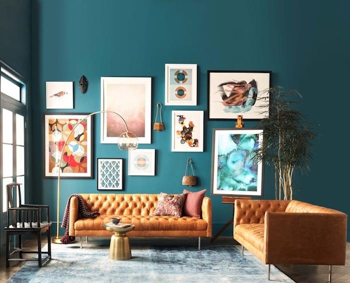 šiuolaikiška alyvinės mėlynos spalvos svetainės tapyba, originalių spalvų rėmų sienų apdaila, oranžinė odinė sofa, mėlynas ir baltas kilimas, auksinis kavos staliukas