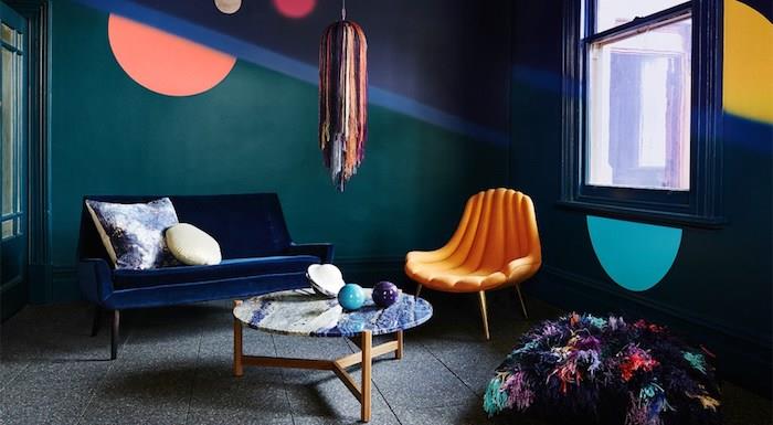 naftos mėlynos svetainės pavyzdys, naftos mėlyni dažai, tamsiai mėlyna sofa, oranžinis fotelis, pilka grindų danga, kavos staliukas su marmurine viršuje, spalvotas osmanas ir pakaba