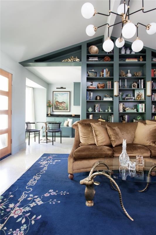 oturma odasında ördek mavisi bir dekorasyon fikri, kahverengi kadife kanepeye uyan küçük pirinç dokunuşlarla ördek mavisi bir kitaplık duvarı