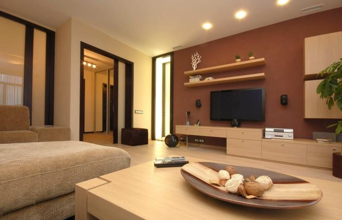 didelis šviesaus medžio stalas, rudi sienų dažai, lentynos, televizoriaus spintelė, pakabinami elementai, didelė smėlio spalvos sofa