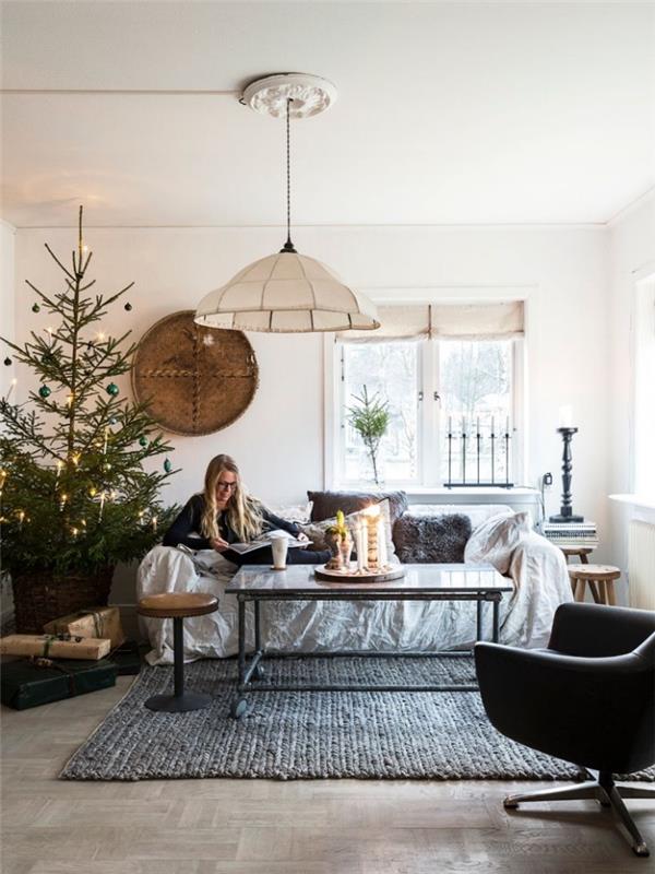 Skandinaviškas ir tradicinis atostogų dvasios dekoras, poilsio zona gyvenamajame kambaryje su nedidele sofa, papildyta pilkomis dirbtinio kailio pagalvėlėmis, ir prie jos pastatyta Kalėdų eglutė