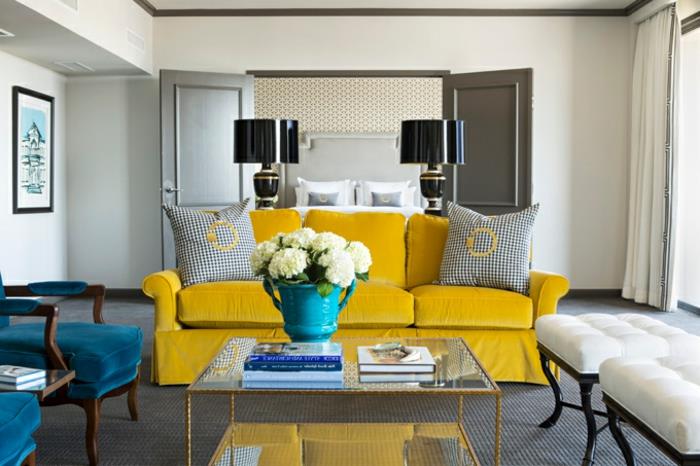 çağdaş bir oturma odasında mavi ve sarı dekor, ördek mavisi sandalyeler, sarı kanepe, sehpa, gri halı, beyaz minderli ahşap tabureler, şık siyah lambalar