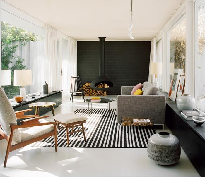 moderni svetainė su kaimiškomis savybėmis, juodos spalvos svetainės apdaila, skandinaviški baldai, aptakiai pilka sofa, pakabinamas židinys, dryžuotas kilimas, langai nuo grindų iki lubų