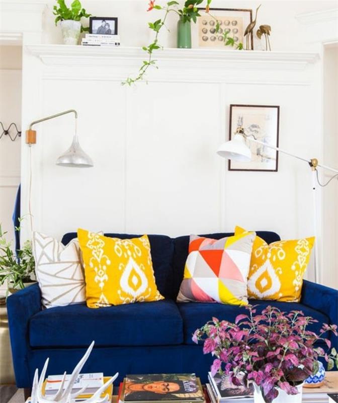 İskandinav oturma odası, lacivert kanepe, sarı ve rengarenk minderler, beyaz duvar, duvar rafı, yeşil bitkiler, dergi ve sanat kitapları yığınları, sarı ve mavi bohem dekorasyonu nasıl düzenleneceğine dair fikir
