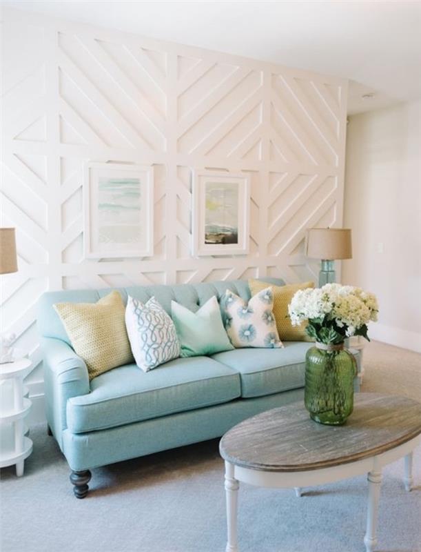 vandens žalia sofa, mėlynos, baltos ir geltonos pagalvėlės, medinis kavos staliukas, pilkas kilimas, balta sienų apdaila, žalia gėlių vaza su puokšte