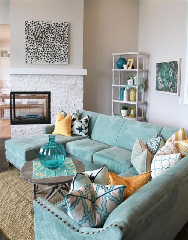 vandens žalios spalvos sofos idėja modernioje pajūrio svetainėje, apvalus medinis kavos staliukas, smėlio spalvos kilimas, akmeninis židinys, perlų pilka spalva, mėlynos, baltos ir geltonos pagalvėlės
