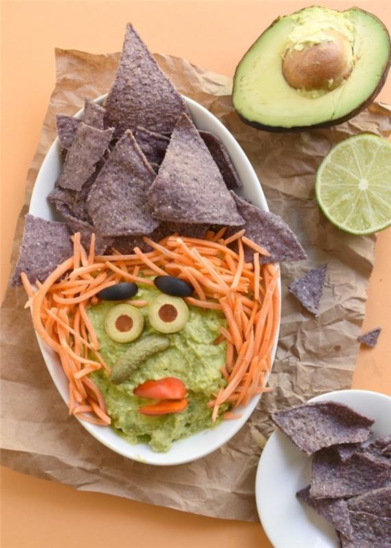 guacamole za aperitiv za noč čarovnic v obliki čarovniške glave, okrašen z ajdovimi tortiljinimi čipsi, olivami, naribanim korenjem in mini kumaricami