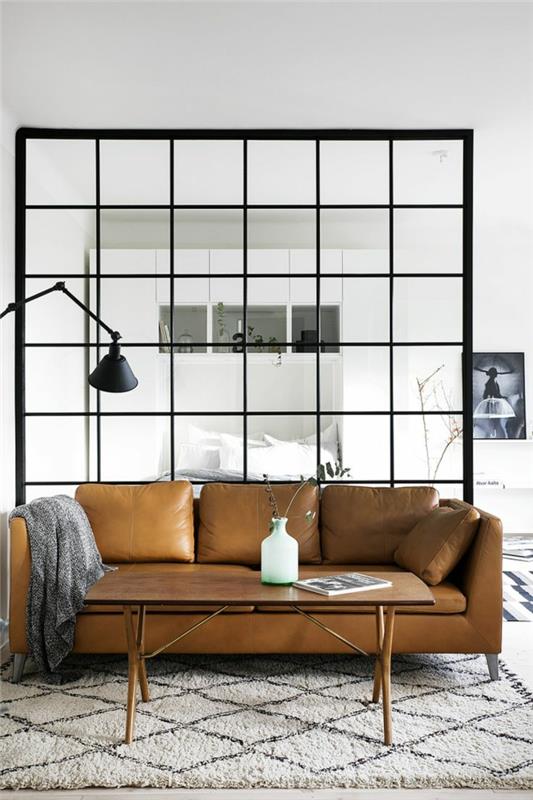 svetainė ir miegamasis su stiklo pertvara, ruda odinė sofa, stačiakampis kavos staliukas, juoda sulankstoma šviesa pramoniniu stiliumi