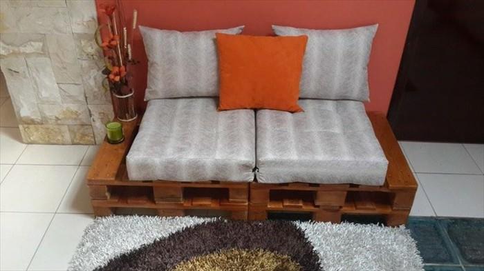 mažas sofas dviem žmonėms, pagamintas iš padėklų, nudažytų ruda spalva, padengtas dryžuotomis pilkomis pagalvėmis ir dekoruotas maža oranžine pagalve, padėklų kušete
