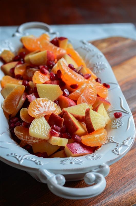 šviežių vaisių salotos, mandarinai, obuoliai, granatų sėklos, balta lėkštė su ornamentais