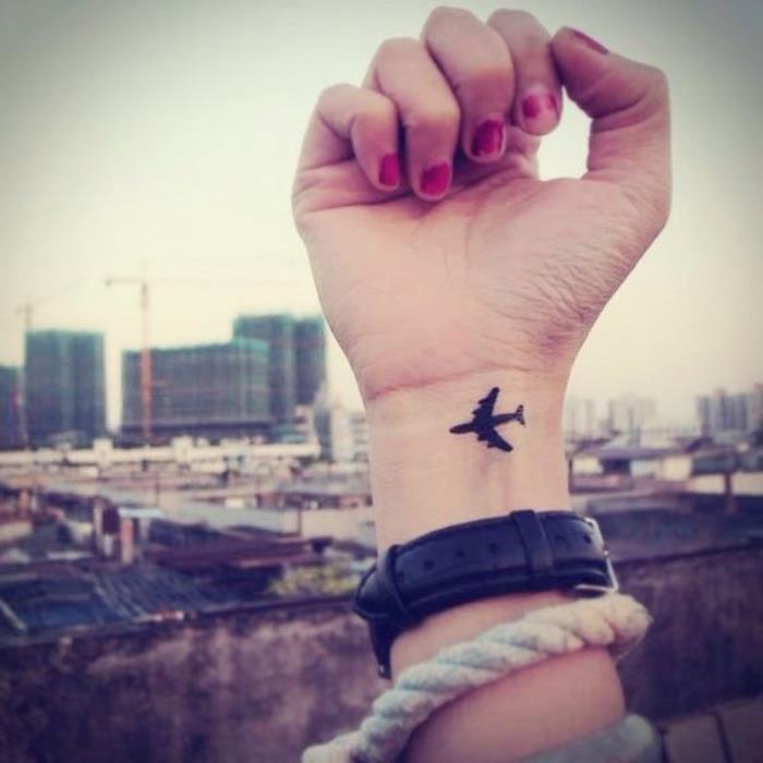 maža juoda lėktuvo riešo tatuiruotė, maža dilbio tatuiruotė, miesto kraštovaizdis fone