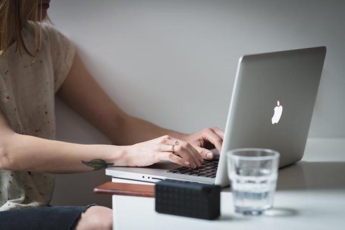 moteris sėdi, spausdina „MacBook“, palieka riešo tatuiruotę, mažą lotoso gėlės tatuiruotę