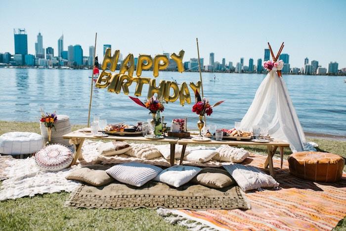 gimtadienio lauko puošmena lauke, prašmatni bohemiška gimtadienio puošmena su žemu mediniu stalu, apsuptu pagalvėlių, auksiniais balionais ir balta palapine šalia