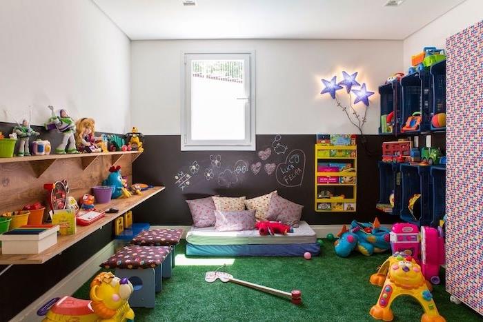 šiferiu dažytos pagrindinės sienos sekcija su vaikų piešiniais, lentynomis ir plastikinėmis dėžėmis, žolės kilimėlių imitacijomis, vaikų žaislais