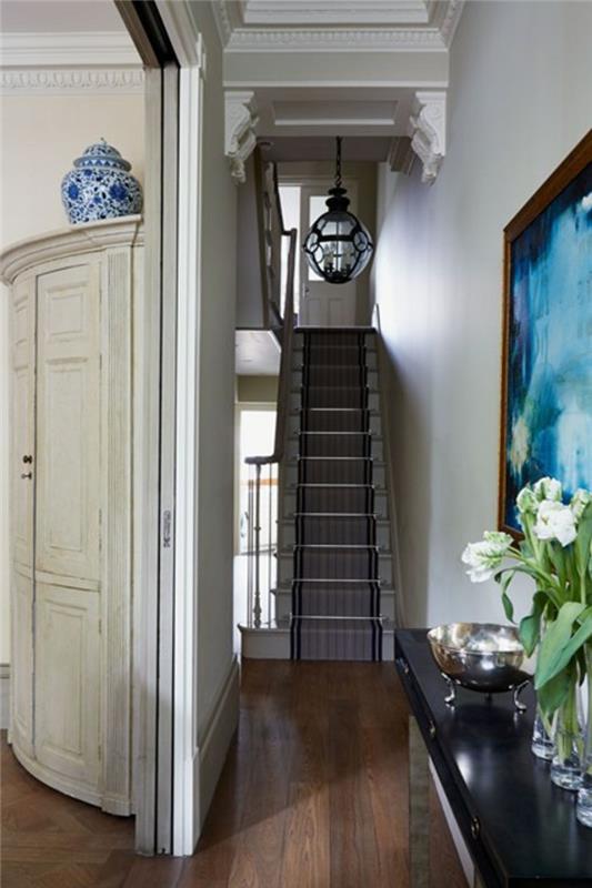 ozke stopnice s črtasto preprogo, temna lesena tla, stene z mavčnimi detajli, slika v modrem okvirju, blizu mize s tulipani v vazah, dekor na hodniku