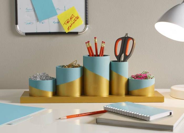 žingsnio tėčio dovanų idėjos, šviesiai mėlynos ir auksinės spalvos biuro reikmenų organizatorius su smeigtukais, guminėmis juostelėmis, pieštukais, žirklėmis ir sąvaržėlėmis ant biuro stalo su užrašų knygelėmis ir lempa