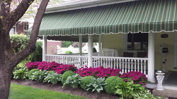 veranda su turėklais ir medinėmis kolonomis, nudažyta balta spalva, motorizuota tenta žalios spalvos žalios spalvos ir baltos ir geltonos spalvos smulkiomis juostelėmis, sodas su fuksijos spalvos gėlėmis ir žaliais rutuliais