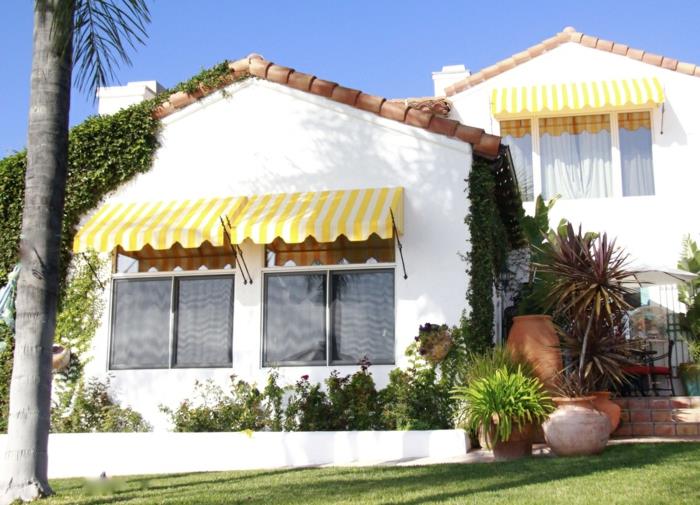 priemiesčio namas su baltomis sienomis, tentu iš geltono audinio ir baltomis vertikaliomis juostelėmis, lauko markizė, saulės markizė, žalia veja priešais namą