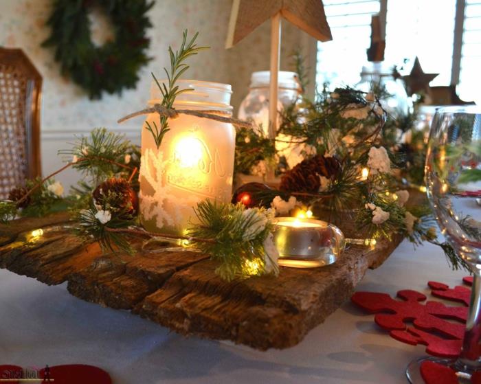 nepermatomi stikliniai indeliai su uždegtomis žvakėmis, neapdorotas medinis valgomojo stalas, girlianda mažose lemputėse, raudonos snaigės, kalėdinis vainikas iš natūralių šakų