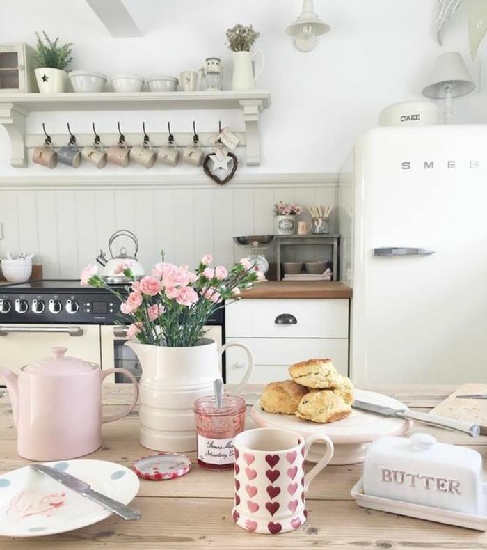 primer elegantne podeželske mize v lesu, bela kuhinja in sprednja stran hladilnika, posoda in skodelice, okras rožnatih cvetov
