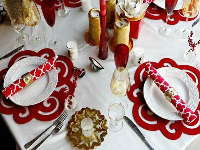 Kalėdų stalo puošmena, baltos lėkštės, raudonos kilimėliai, auksinės žvakidės, balta staltiesė