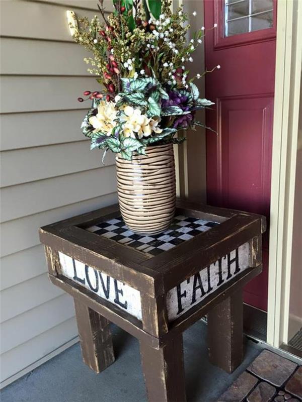 priešais įėjimą į namą sumontuotas nedidelis sodo stalas, paletiniai sodo baldai, kačių dryžuota vaza su sodo gėlėmis