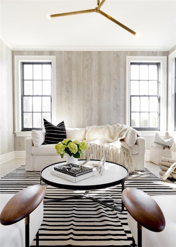 jauki svetainės apdaila su baltomis lubomis ir grindimis, šviesiai pilka lentų medžio sienų apdaila, baltos ir juodos spalvos dryžuotas kilimas