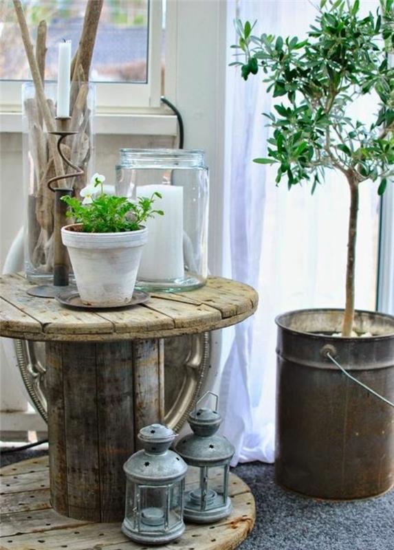okras iz lesenega bobna, miza iz surovega lesa, starinske luči, cvetlični lonec iz beljene terakote, stekleni kozarec s svečo v notranjosti, drevo posajeno v kovinsko vedro
