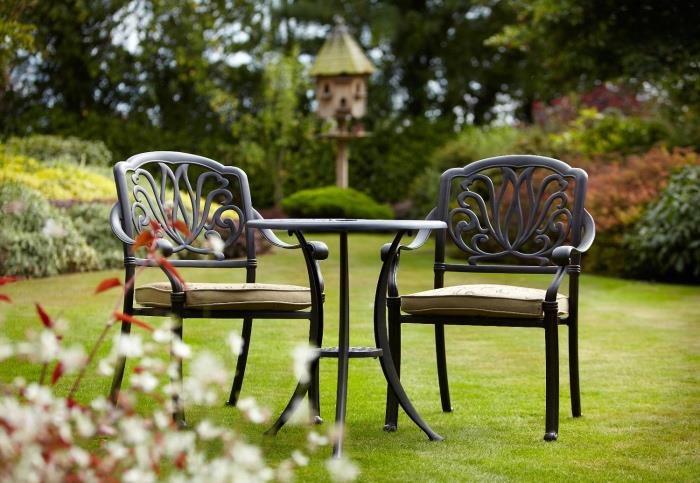 paprasti sodo baldai ant vejos, juodas apvalus stalas ir juodos kėdės su smėlio spalvos sėdynių pagalvėlėmis, keli krūmai ir medžiai pasienyje