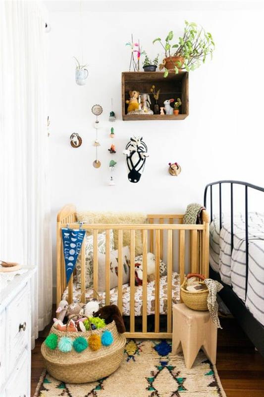 kūdikio mergaitės kambario apdaila, šviesaus medžio lova, kvadrato formos spintelės lentyna, nedidelė kvadrato formos taburetė, vazonai su žaliais augalais, maži dekoratyviniai daiktai, kabantys ant sienos, stačiakampis kilimas etniniu stiliumi