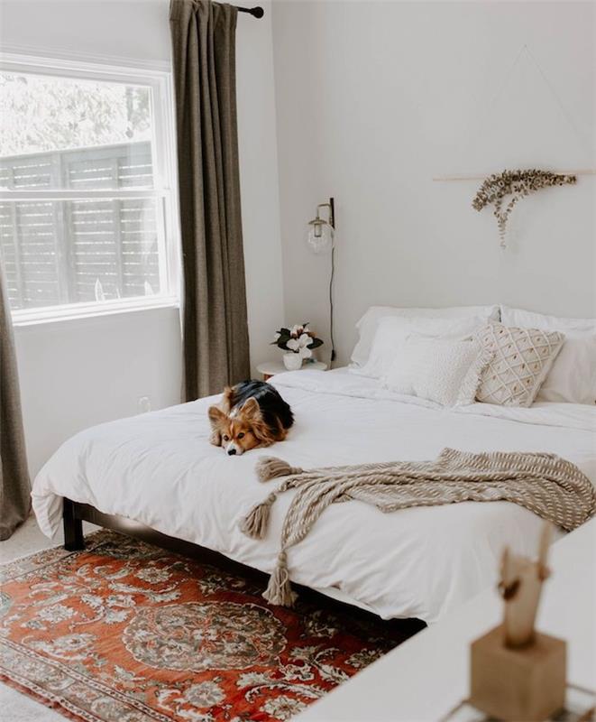 prašmatnus bohemiškas kilimas kambaryje su baltomis sienomis, baltu namų skalbiniu, mielu šunimi, pilka lovos užtiesalu, šiaurietišku dekoru