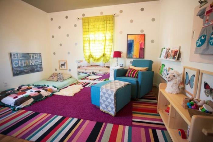 balta siena, pilki taškeliai, knygų lentynos, įvairiaspalviai kilimėliai, „blei“ fotelis, „Montessori“ kūdikio lova ant grindų, bohemiškas stilius, žaislai, dekoracijos