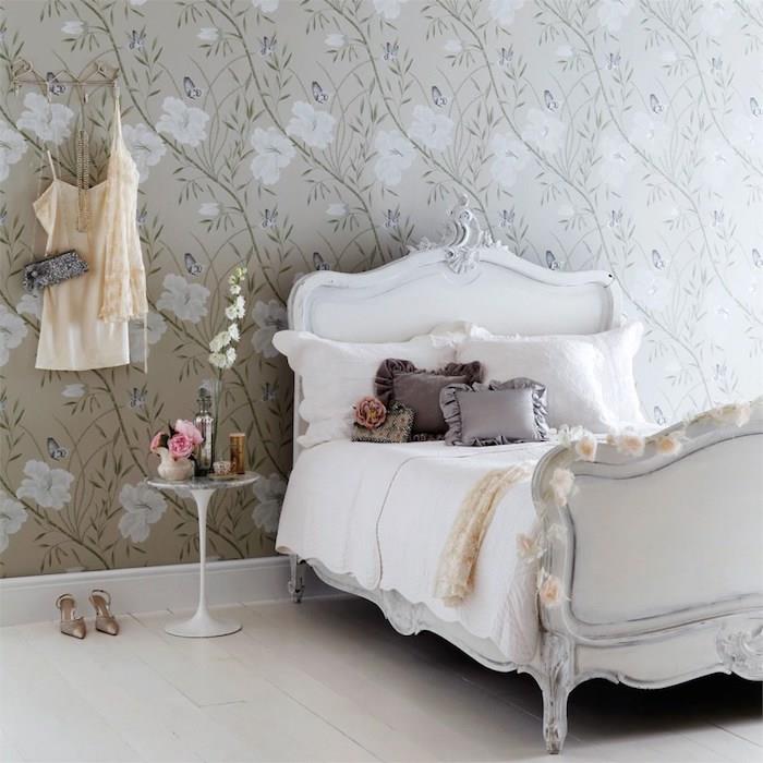 Kadınsı tasarımlı bir yatak odasında eski duvar kağıdı ve barok yatak, beyaz keten üzerinde gri minderler, ağartılmış parke, gri bir arka plan üzerinde çiçek motifli duvar kaplaması