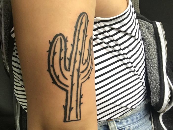 moters rankos tatuiruotė, stilizuoto dizaino juodo kaktuso tatuiruotė, mėlyni džinsai ir dryžuota palaidinė
