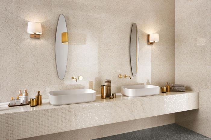 kopalniški dekor v bež in belih odtenkih, stenska obloga iz bež ploščic, model ovalnega ogledala in bakreni kopalniški dodatki