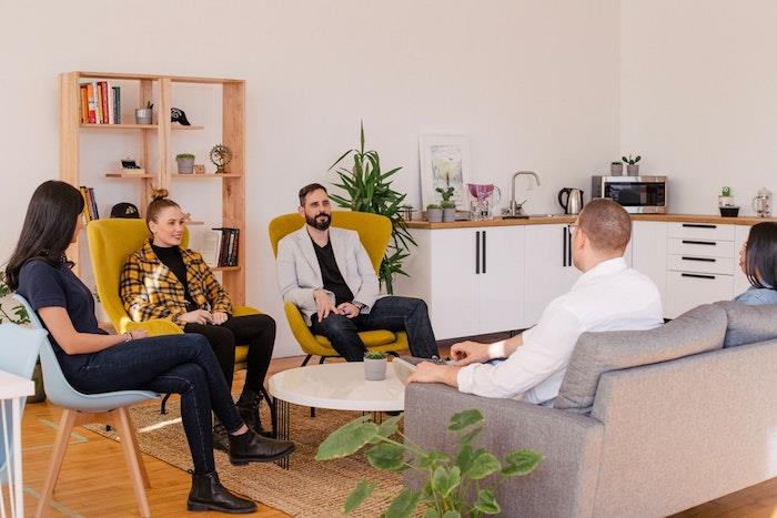 Pilka sofa, mažas apvalus kavos staliukas, medinė lentyna, madinga biuro išdėstymo idėja