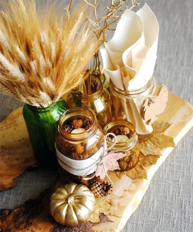 jesenska dekoracija mize, osrednji del lesene deske s steklenimi kozarci, napolnjenimi z borovimi storžki, vaza s pšeničnimi klasovi in ​​zlatimi bučami
