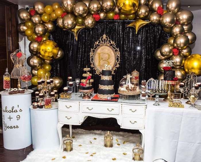 30 -ojo gimtadienio temos pavyzdys, auksinių balionų arka, juoda uždanga, keksiukas, pyragas ir maži auksiniai bei juodi skanėstai