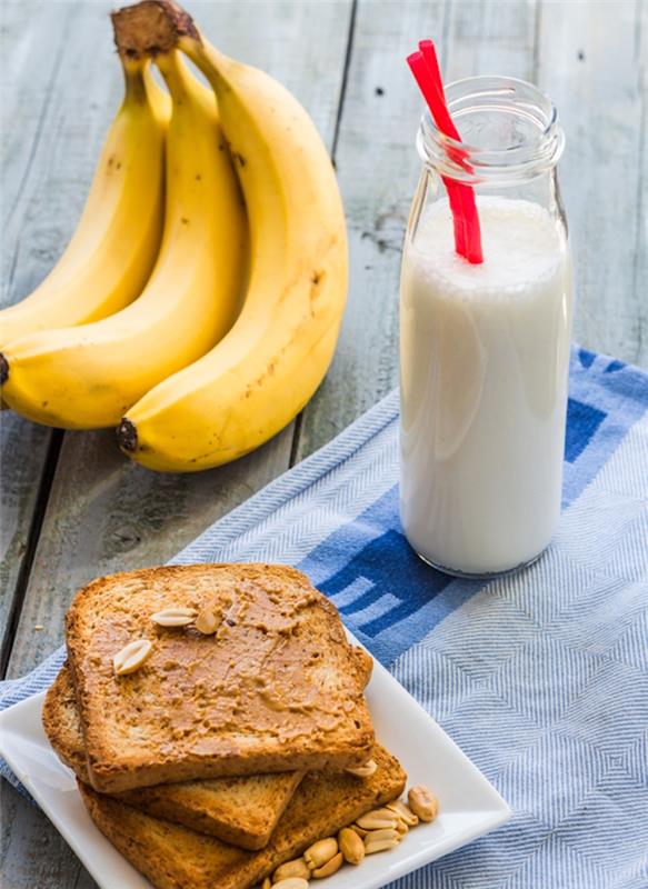 žemės riešutų sviesto skrebučiai su žemės riešutais ir nedidelis stiklinis indelis, pripildytas pieno, bananų