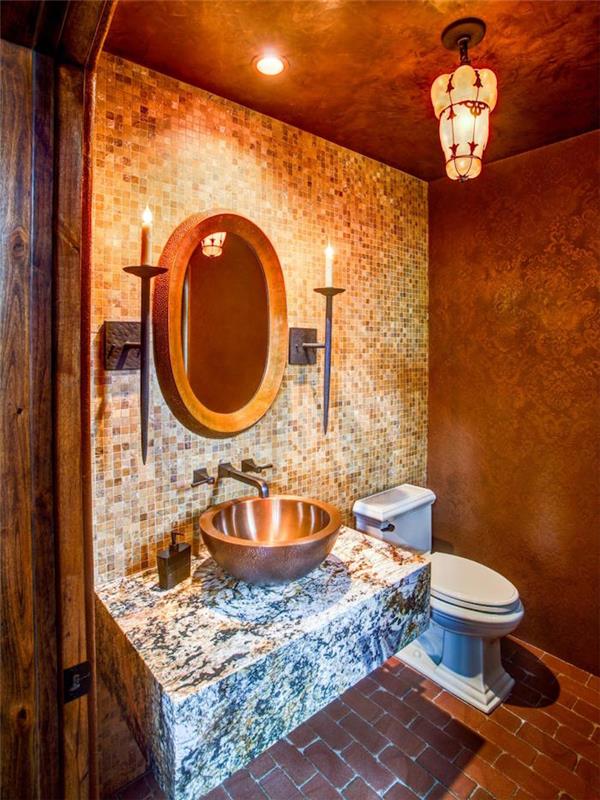 ideje za majhno kopalnico, stranišče z rjavimi stenami, eno z mozaičnimi ploščicami, v različnih odtenkih rjave barve, drugo z rjavo barvo in baročnim vzorcem, velika marmorna plošča s kovinskim umivalnikom, ovalno ogledalo z lesenim okvirjem, dve prižgani sveči v držalih