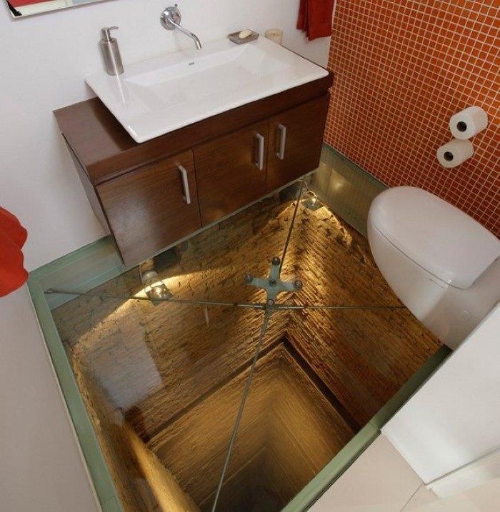 prenova kopalnice, majhno stranišče z dvema belima stenama, eno steno, prekrito z oranžnim mozaikom, rjava lesena omara z ravnim belim umivalnikom, bela straniščna školjka, pogled skozi steklena tla, ki razkriva globok tunel spodaj